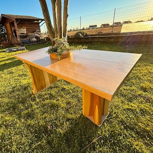 Può includere: Tavolo in legno fatto a mano con finitura marrone chiaro. Il tavolo ha un piano rettangolare e un design unico delle gambe. Il tavolo è all'aperto su un prato. Le dimensioni del tavolo sono di circa 150 cm di lunghezza e 75 cm di larghezza.