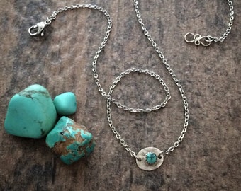 May include: A silver chain necklace with a turquoise stone pendant. The pendant is oval shaped and has a hammered texture. The necklace is on a wooden surface with turquoise stones.
