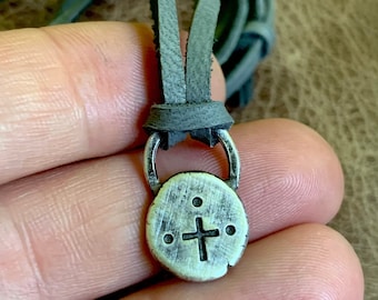 Puede incluir: Un collar con un colgante de plata redondo con una cruz y tres puntos. El collar está en un cordón de cuero gris.