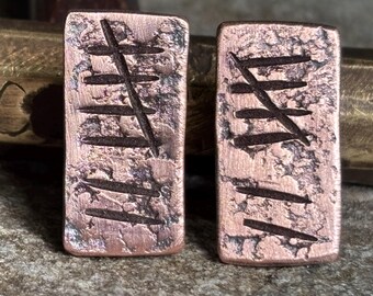 May include: Two rectangular copper pendants with etched symbols. The pendants have a textured surface and a dark patina. The symbols appear to be a series of lines and cross-hatching. The background includes a brass object and a stone surface.