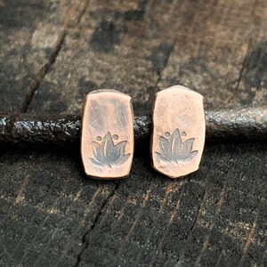 May include: A pair of copper earrings with a lotus flower design. The earrings are rectangular in shape and have a rustic finish.