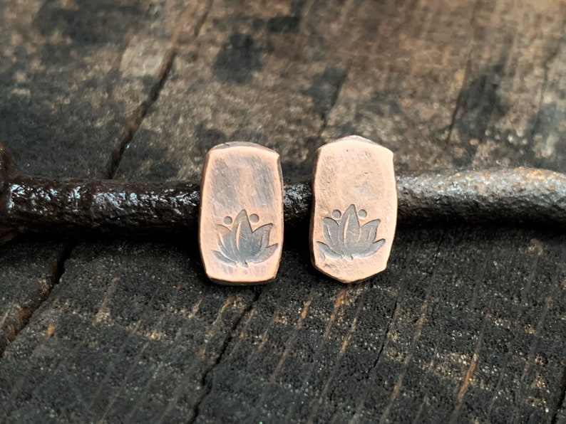 May include: A pair of copper earrings with a lotus flower design. The earrings are rectangular in shape and have a rustic finish.