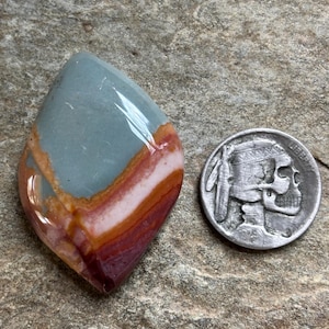May include: A polished gemstone with a mix of colors, including blue-green, pink, and deep red-brown, is displayed next to a silver coin. The gemstone has a smooth, glossy surface and a unique, layered appearance. The coin features a Native American profile.