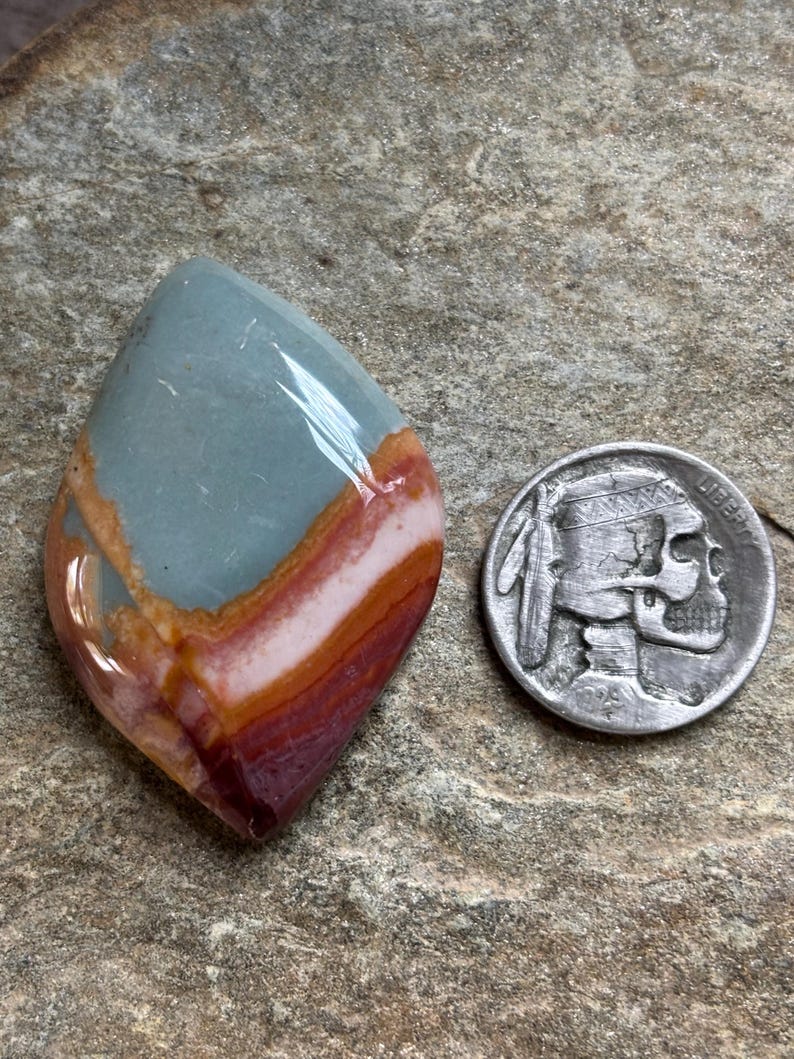 May include: A polished gemstone with a mix of colors, including blue-green, pink, and deep red-brown, is displayed next to a silver coin. The gemstone has a smooth, glossy surface and a unique, layered appearance. The coin features a Native American profile.
