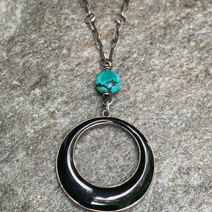 May include: A silver-toned necklace with a large black circular pendant. The necklace features a turquoise bead and a chain. The pendant is a glossy black with a silver border. The necklace is displayed on a gray stone surface.