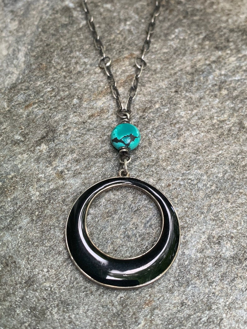 May include: A silver-toned necklace with a large black circular pendant. The necklace features a turquoise bead and a chain. The pendant is a glossy black with a silver border. The necklace is displayed on a gray stone surface.