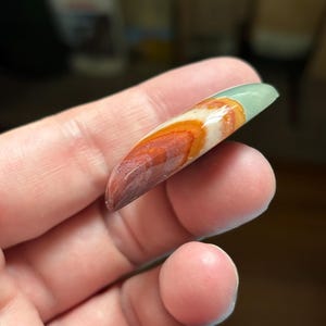 May include: A polished, elongated gemstone with a gradient of colors. The stone transitions from a deep reddish-brown at the tip, through orange and white, to a pale green at the top. The stone is held in a hand.