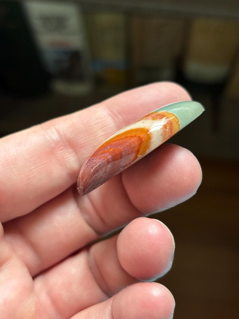 May include: A polished, elongated gemstone with a gradient of colors. The stone transitions from a deep reddish-brown at the tip, through orange and white, to a pale green at the top. The stone is held in a hand.