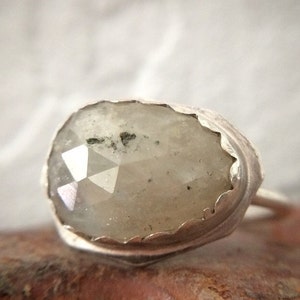 May include: A silver ring with a large, oval-shaped, grey, faceted gemstone. The gemstone has a rough, natural texture and is set in a silver band with a scalloped edge.