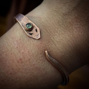 May include: A copper bracelet with a green gemstone set in the center. The bracelet is open and has a curved shape.