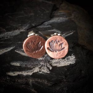 May include: A pair of copper stud earrings with a lotus flower design. The earrings are round and have a silver backing.