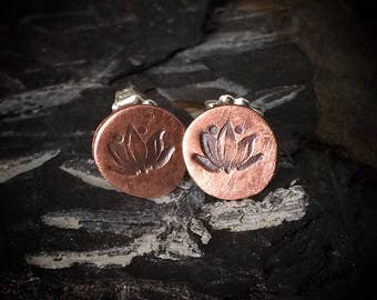 May include: A pair of copper stud earrings with a lotus flower design. The earrings are round and have a silver backing.
