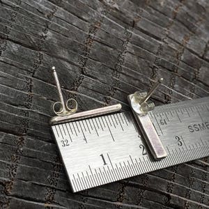 May include: A pair of silver bar earrings measuring approximately 2.5 cm in length. The earrings are shown next to a ruler for size reference.