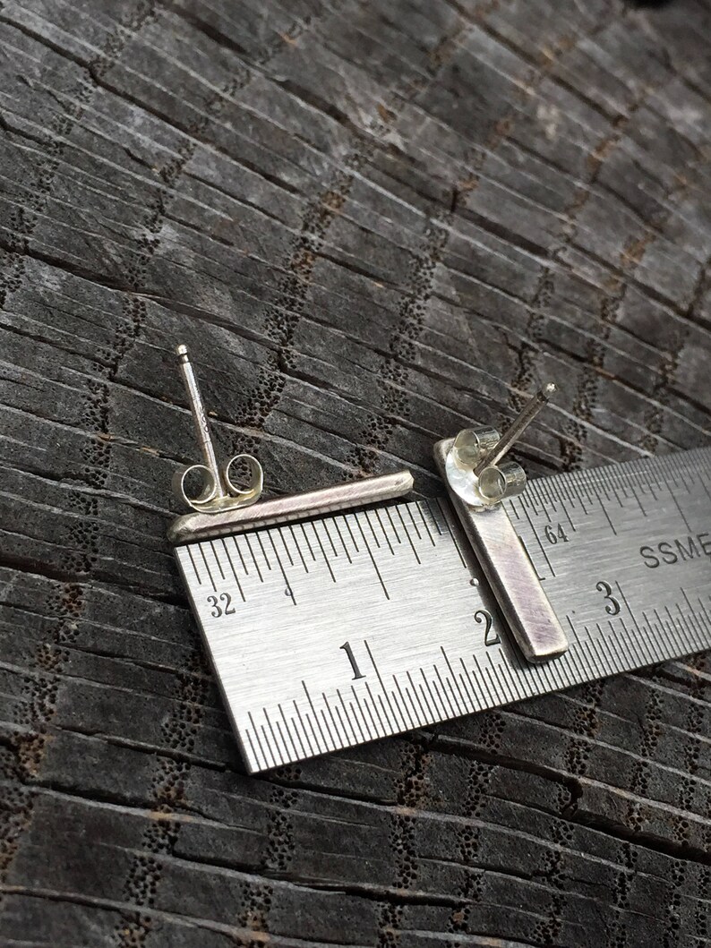 May include: A pair of silver bar earrings measuring approximately 2.5 cm in length. The earrings are shown next to a ruler for size reference.