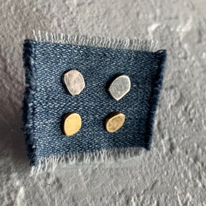 May include: Four small, irregular-shaped metal earrings in silver, gold, and rose gold. The earrings are displayed on a piece of blue denim fabric.