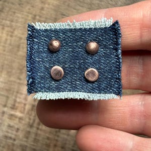 May include: A small, rectangular denim patch with four copper-colored rivets. The denim is a dark blue with a frayed, light blue edge. The rivets are evenly spaced across the patch.