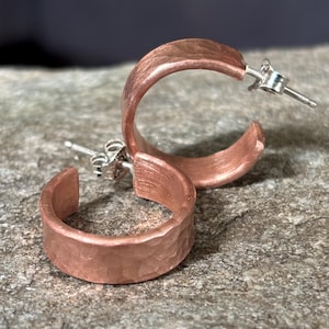 May include: A pair of copper-colored hoop earrings with a hammered texture. The earrings have a post and butterfly backing. The earrings are displayed on a textured, gray surface.