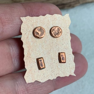 May include: A set of four copper-coloured stud earrings on a tan card. The earrings include two round studs with textured designs and two rectangular studs. The earrings are displayed against a neutral background.