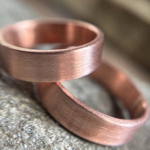 May include: Two copper rings with a brushed metal finish. The rings are a warm, reddish-brown color and have a simple, classic design. The rings are likely wedding bands or promise rings.