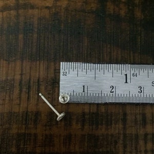 May include: A silver stud earring with a small round post and a flat, circular back. The earring is measured against a ruler, showing it is less than 1 inch in length.