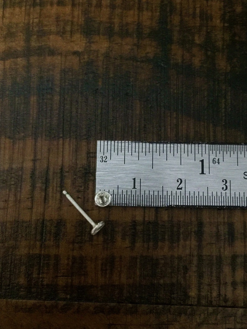 May include: A silver stud earring with a small round post and a flat, circular back. The earring is measured against a ruler, showing it is less than 1 inch in length.