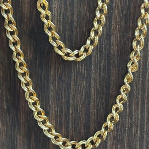 May include: A gold-colored chain necklace with a double-link design. The necklace is displayed against a dark brown wooden background. The chain links are closely connected, creating a textured appearance. The necklace is a jewelry item.
