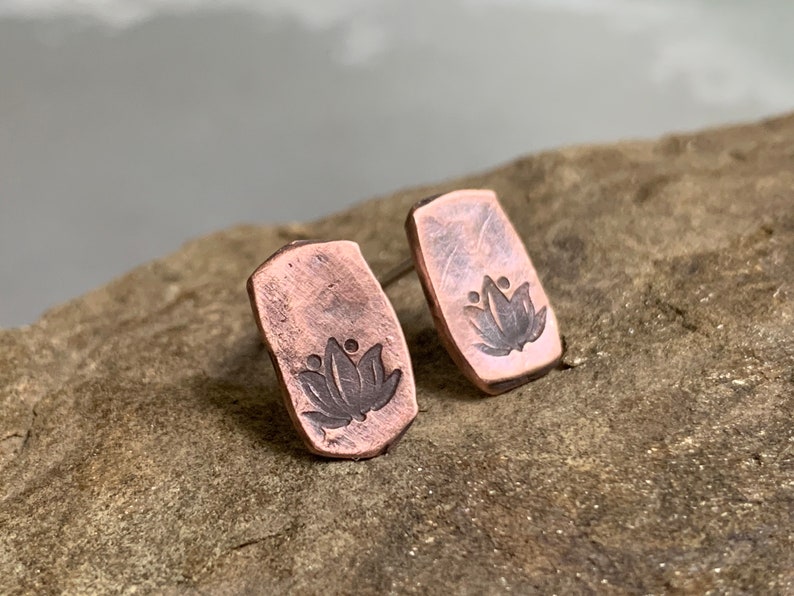 May include: A pair of copper earrings with a lotus flower design. The earrings are a rectangular shape and have a hammered texture.
