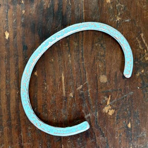 May include: A turquoise-colored metal cuff bracelet with a textured surface. The open-ended bracelet has a distressed, vintage appearance, with a copper-colored edge. The bracelet is set against a dark wood background.