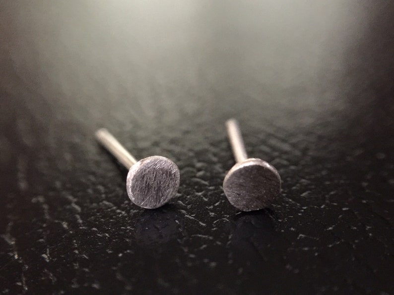 May include: A pair of silver stud earrings with a circular design. The earrings are on a dark surface.
