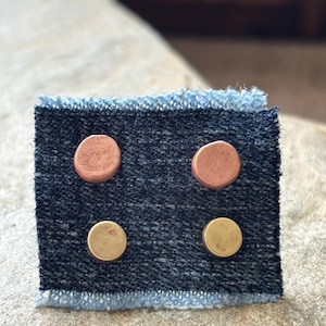 May include: Two pairs of circular stud earrings attached to a square piece of dark blue denim fabric with frayed light blue edges. The top pair is made of copper, while the bottom pair is made of brass, both featuring a smooth, flat surface.