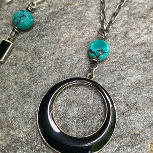 May include: A silver necklace with a large black circular pendant and a turquoise bead. A matching earring with a turquoise bead and a black rectangular accent is also visible. The jewelry is displayed on a gray stone surface.