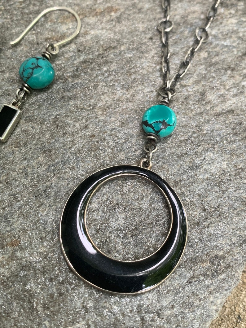 May include: A silver necklace with a large black circular pendant and a turquoise bead. A matching earring with a turquoise bead and a black rectangular accent is also visible. The jewelry is displayed on a gray stone surface.