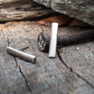 May include: Two silver bar earrings on a rustic wooden surface. The earrings are simple and minimalist, with a rectangular shape.