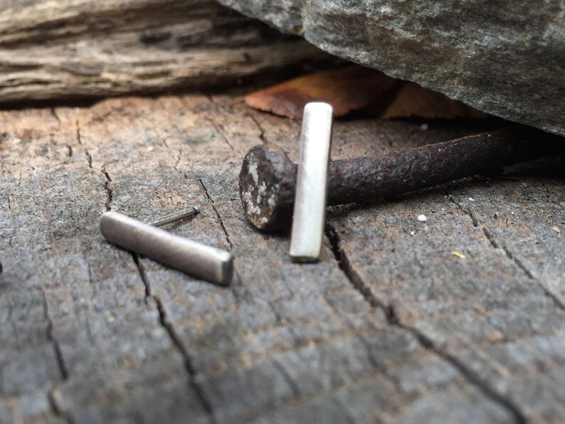 May include: Two silver bar earrings on a rustic wooden surface. The earrings are simple and minimalist, with a rectangular shape.