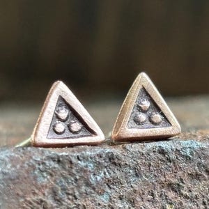 May include: A pair of stud earrings featuring a triangular shape with a textured copper-toned finish. Each earring displays a recessed center containing three small, raised circular dots arranged in a triangular pattern within the frame.