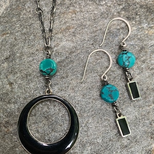 May include: A silver necklace with a large black circle pendant and a turquoise bead. Matching silver earrings with turquoise beads and black rectangular accents complete the jewelry set.