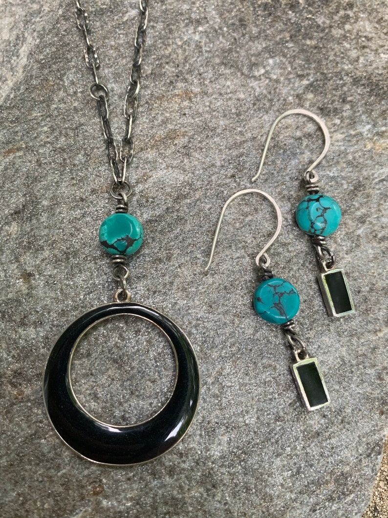 May include: A silver necklace with a large black circle pendant and a turquoise bead. Matching silver earrings with turquoise beads and black rectangular accents complete the jewelry set.