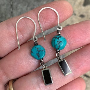 May include: A pair of handcrafted earrings featuring turquoise beads and rectangular black insets. The earrings have silver-colored hooks and wire detailing, showcasing a bohemian style. The turquoise beads are approximately 0.5 inches in diameter.