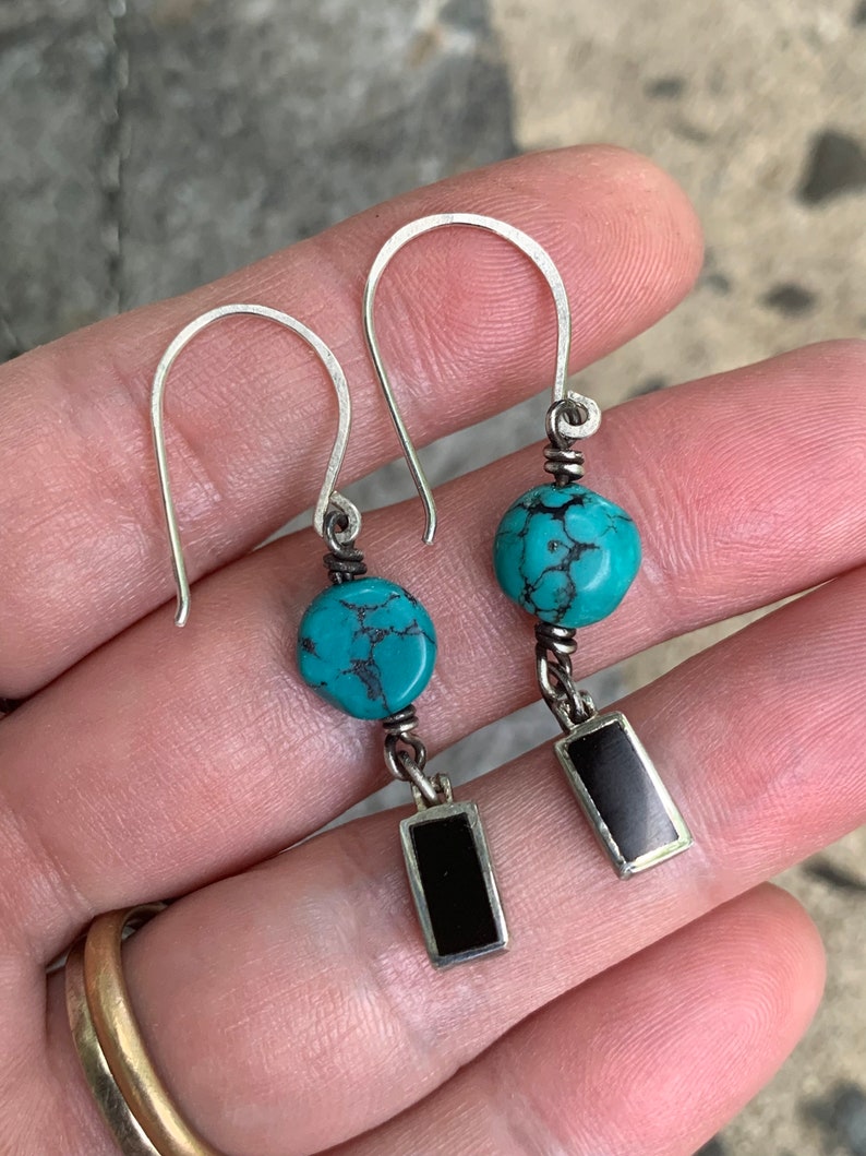May include: A pair of handcrafted earrings featuring turquoise beads and rectangular black insets. The earrings have silver-colored hooks and wire detailing, showcasing a bohemian style. The turquoise beads are approximately 0.5 inches in diameter.