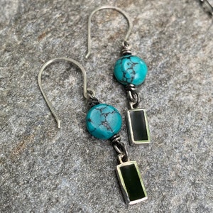 May include: A pair of silver earrings with turquoise beads and black rectangular pendants. The earrings have a hook design and are displayed on a gray, textured surface. The turquoise beads are round and have a natural, mottled appearance.