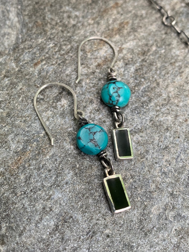 May include: A pair of silver earrings with turquoise beads and black rectangular pendants. The earrings have a hook design and are displayed on a gray, textured surface. The turquoise beads are round and have a natural, mottled appearance.