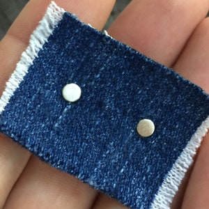 May include: Two small, round, silver stud earrings on a piece of blue denim.