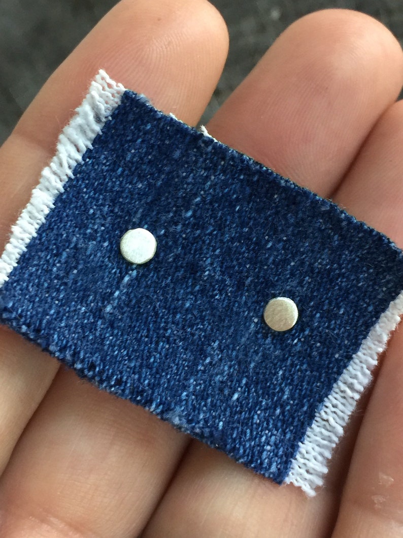 May include: Two small, round, silver stud earrings on a piece of blue denim.