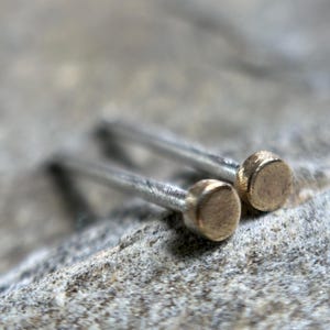 May include: Close-up of two stud earrings. Each earring has a silver post and a small, round, gold-colored head. The earrings are resting on a textured, gray surface. The focus is on the jewelry.