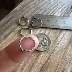 May include: Three silver hoop earrings of different sizes, with the smallest hoop being held by a finger. The hoops are shown next to a ruler with the numbers 2, 3, and 4 visible.