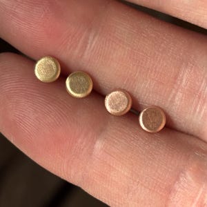 May include: Four small, round stud earrings in varying shades of gold and copper are displayed. The earrings are held in a hand, showcasing their simple, minimalist design. The earrings are likely made of metal and are suitable for everyday wear.