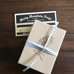 May include: A small, brown box with a white ribbon tied around it. The box has a design of a feather on the top. The box is sitting on a wooden surface. The text on the card reads "Misty Mountain Forge, hand crafted jewelry by artist, metalwork, follow on Instagram, SHOP: www.er"