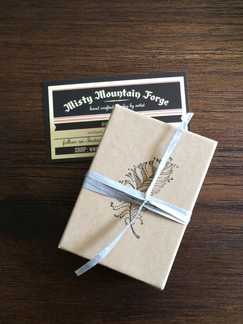 May include: A small, brown box with a white ribbon tied around it. The box has a design of a feather on the top. The box is sitting on a wooden surface. The text on the card reads "Misty Mountain Forge, hand crafted jewelry by artist, metalwork, follow on Instagram, SHOP: www.er"