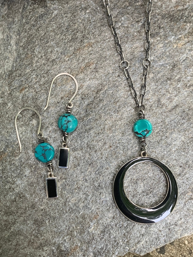 May include: A silver necklace and earring set featuring turquoise beads and black rectangular accents. The necklace has a large, circular pendant with a black interior. The earrings are hook style.