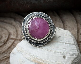 May include: A silver ring with a large, faceted, pink gemstone. The gemstone is set in a textured, circular bezel with a twisted rope design around the edge. The ring is displayed on a white seashell.